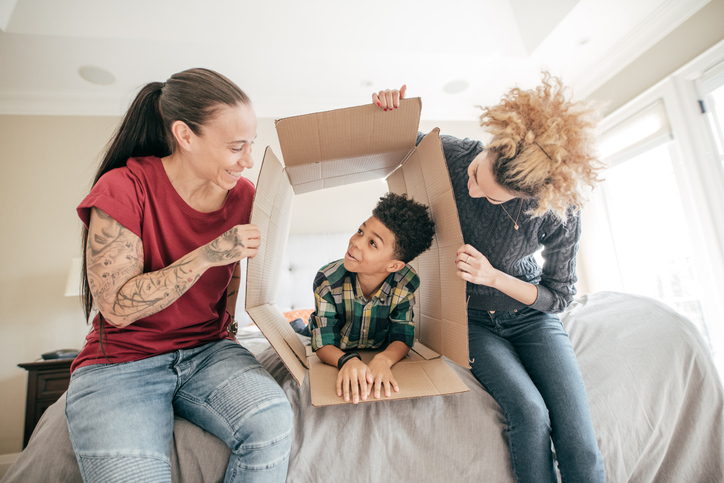 Two women holding out box on bed with little boy playing inside
