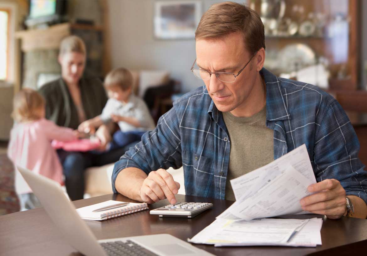 Man using calculator and flipping through papers with family in background